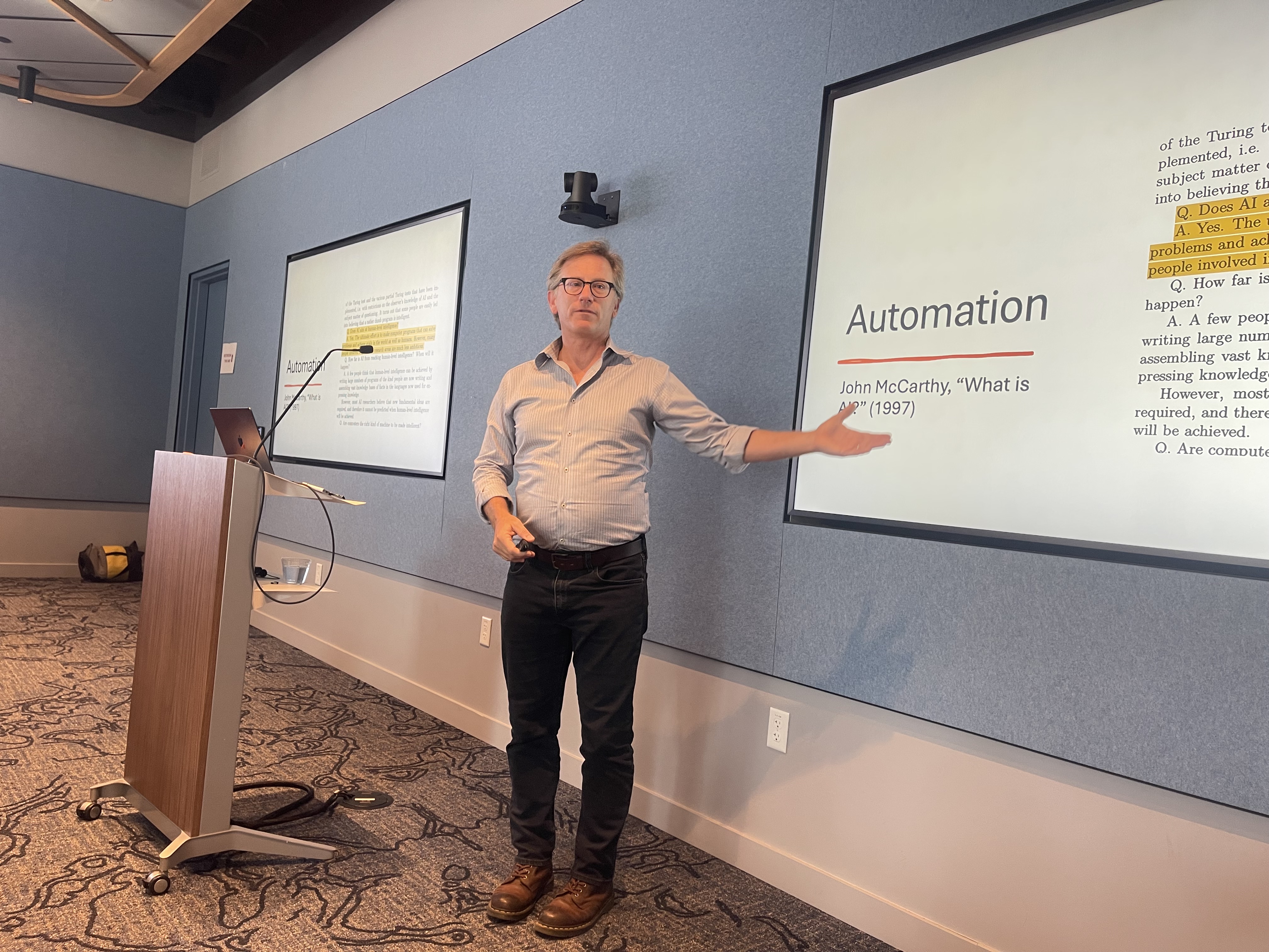 Rob Reich at the Stanford 'AI and the Future of Work' meetup, gesturing toward a slide titled 'Automation' subtitled 'John McCarthy, What is AI? (1997)'.